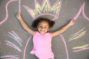 Little African American child lying near chalk drawing of wings and crown on asphalt, top view