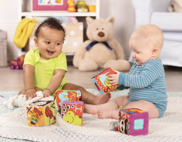 Two smiling babies playing with soft baby blocks on the floor.