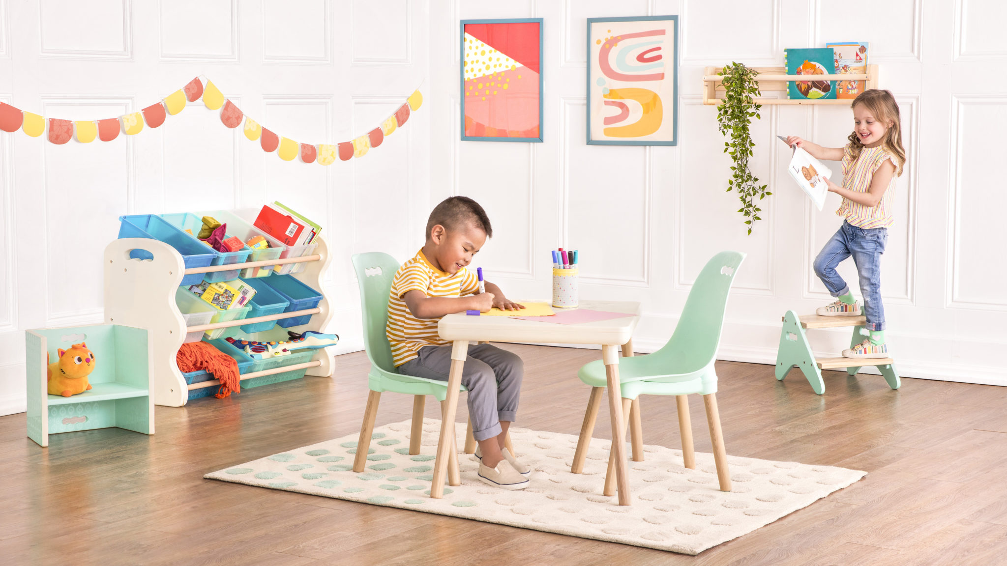 Smiling boy sitting at a kids table and drawing with markers and smiling girl holding a drawing while standing on a kids step stool.