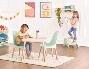 Smiling boy sitting at a kids table and drawing with markers and smiling girl holding a drawing while standing on a kids step stool.