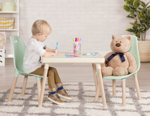 Boy sitting at a kids table with teddy bear across from him while drawing with markers.