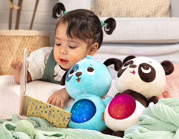 Baby girl reading a board book next to plush toys.